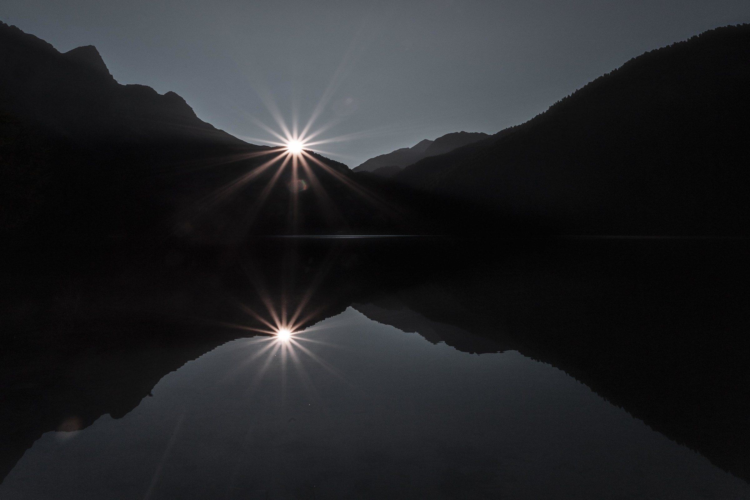 Il sole spunta tra due versanti di montagna e si riflette in un lago scuro.
